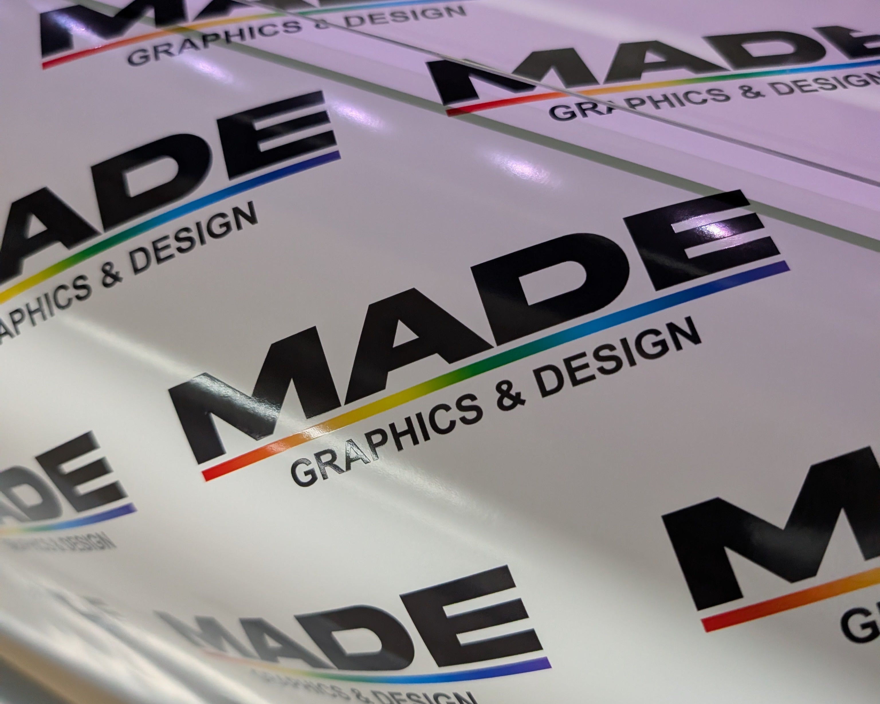 A close-up of a roll of white vinyl or adhesive film printed repeatedly with the logo 'MADE GRAPHICS & DESIGN' in black, with a small rainbow accent line under 'MADE'.